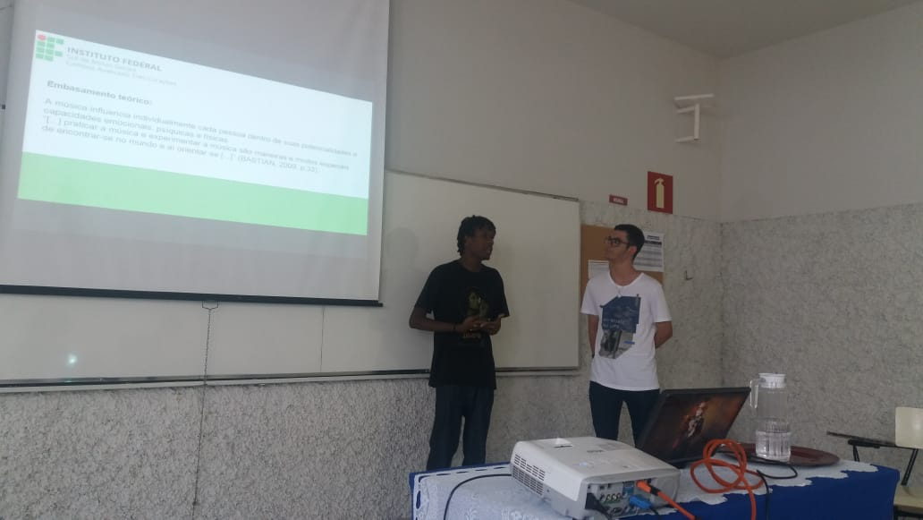 Dois alunos apresentam projeto há um slide projetado na parede sobre o projeto